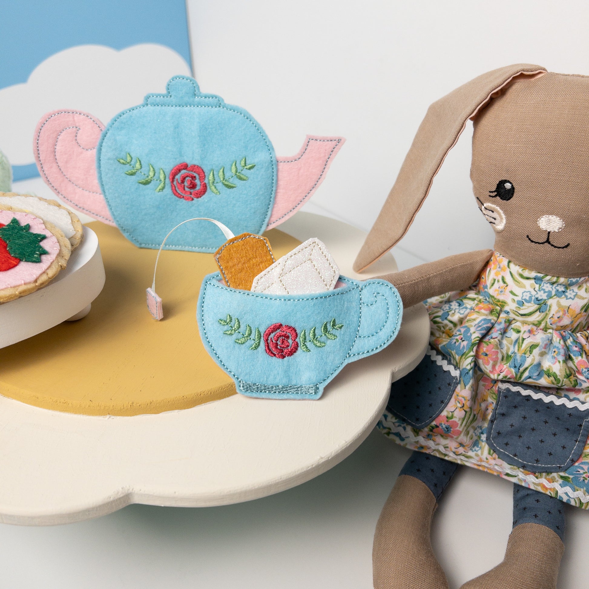 Handmade felt tea set on a cloud-shaped table, including blue teapot and cup with rose embroidery, felt cookies, and tea accessories. A stuffed bunny in a floral dress sits beside the table. Felt cookie matching game visible. Scene set against a background with white clouds on blue sky.