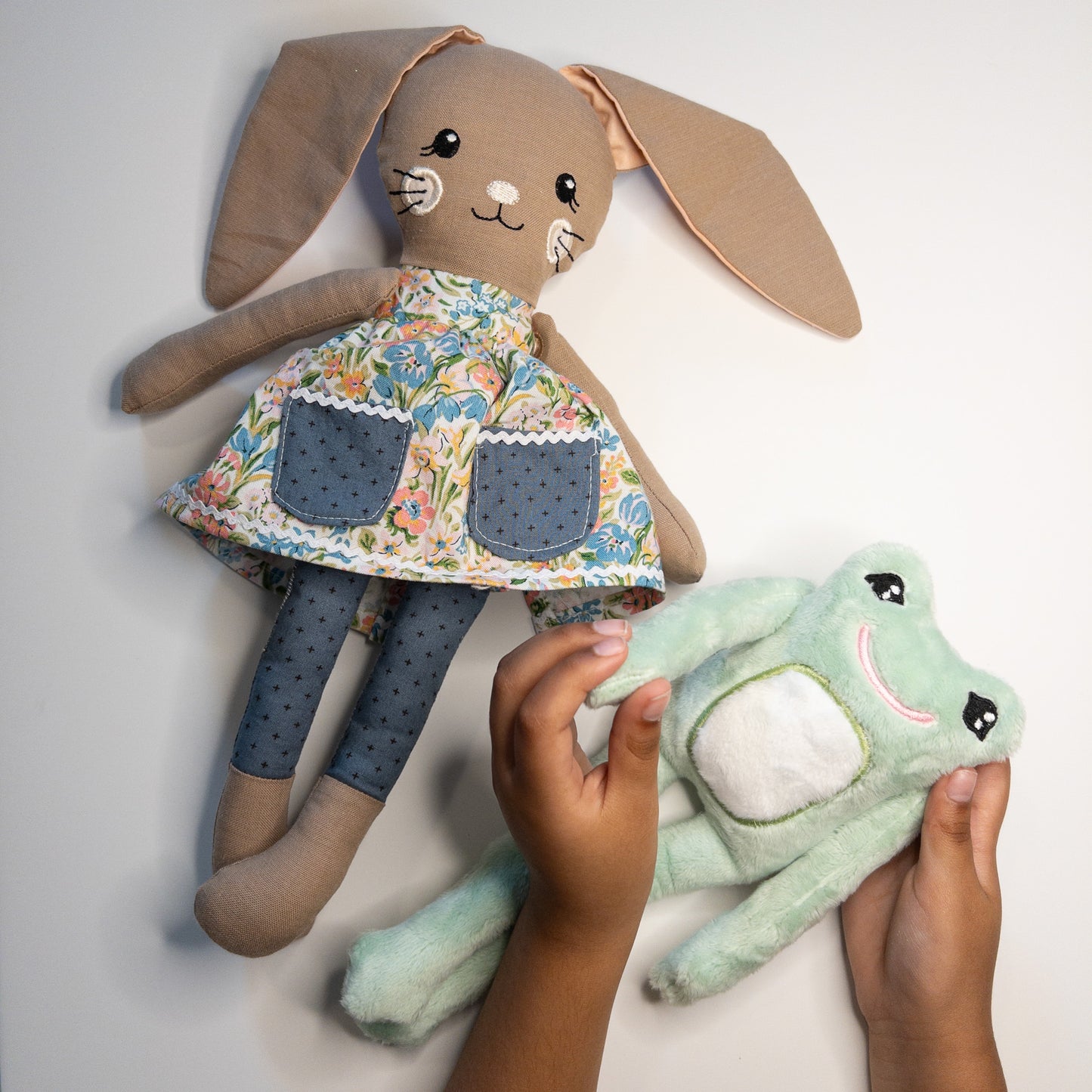 Child's hands playing with handmade stuffed animals - a mint green plush frog and a brown stuffed bunny in a floral dress. The soft, cuddly toys are being held and manipulated, showcasing interactive play with these charming handcrafted companions. Background neutral, focusing on the tactile interaction between child and toys.