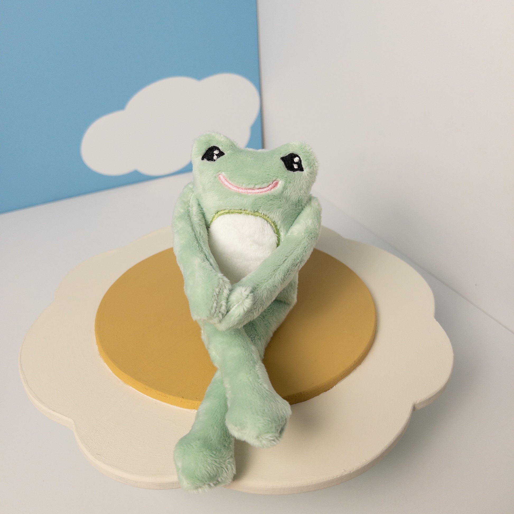 Handmade mint green plush frog stuffed animal sitting with posable legs crossed. Magnetic hands clasped together in front, showcasing interactive features. Frog has friendly embroidered smile and black eyes. Displayed against plain background, highlighting the toy's flexibility and charming pose.