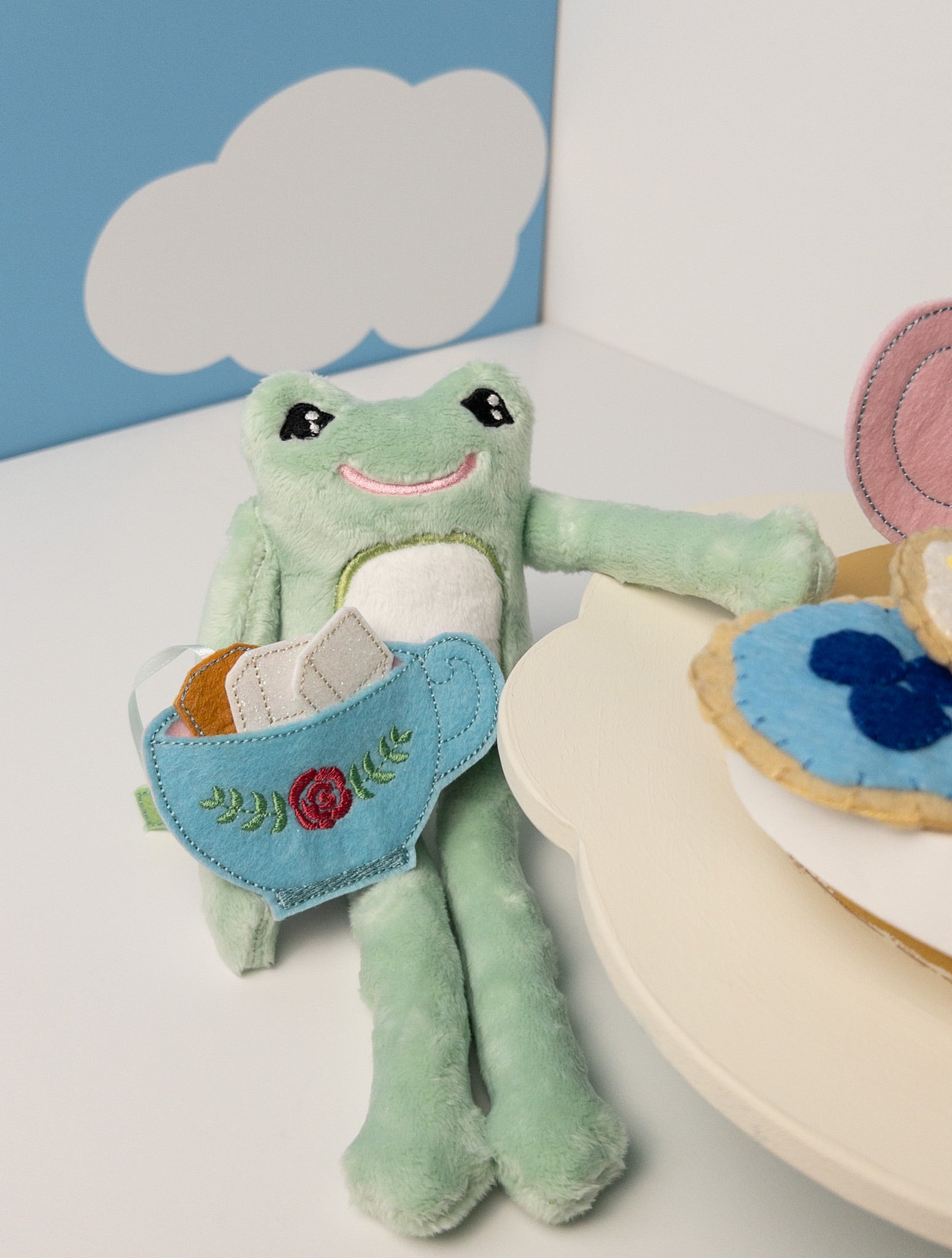 Handmade mint green plush frog stuffed animal holding a blue felt teacup with embroidered rose design. Frog has a friendly embroidered smile and black eyes. Both toys displayed against a light background, showcasing handcrafted details.