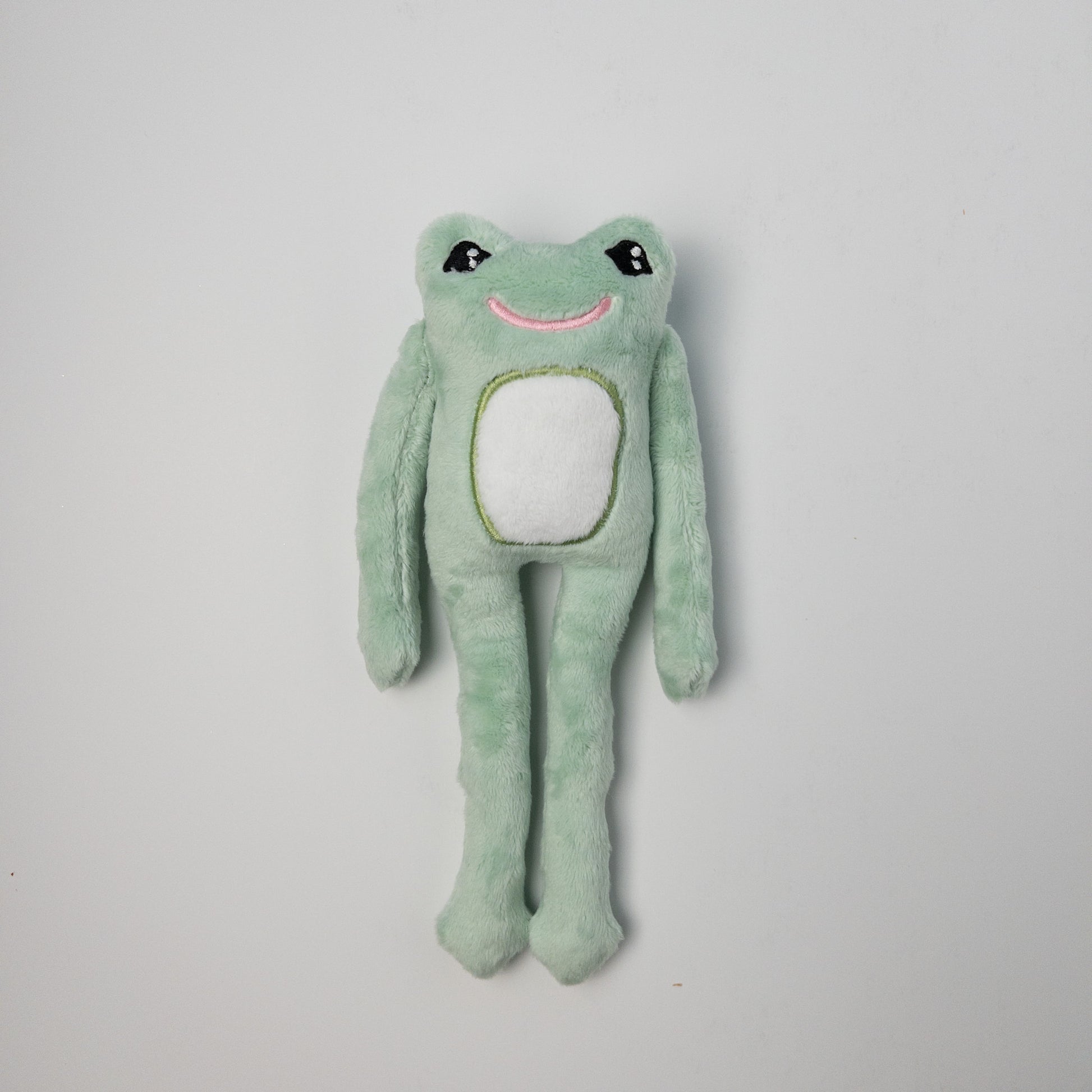 Handmade mint green plush frog stuffed animal with long arms and legs, white belly patch, embroidered smile, and black eyes, positioned against a light background.