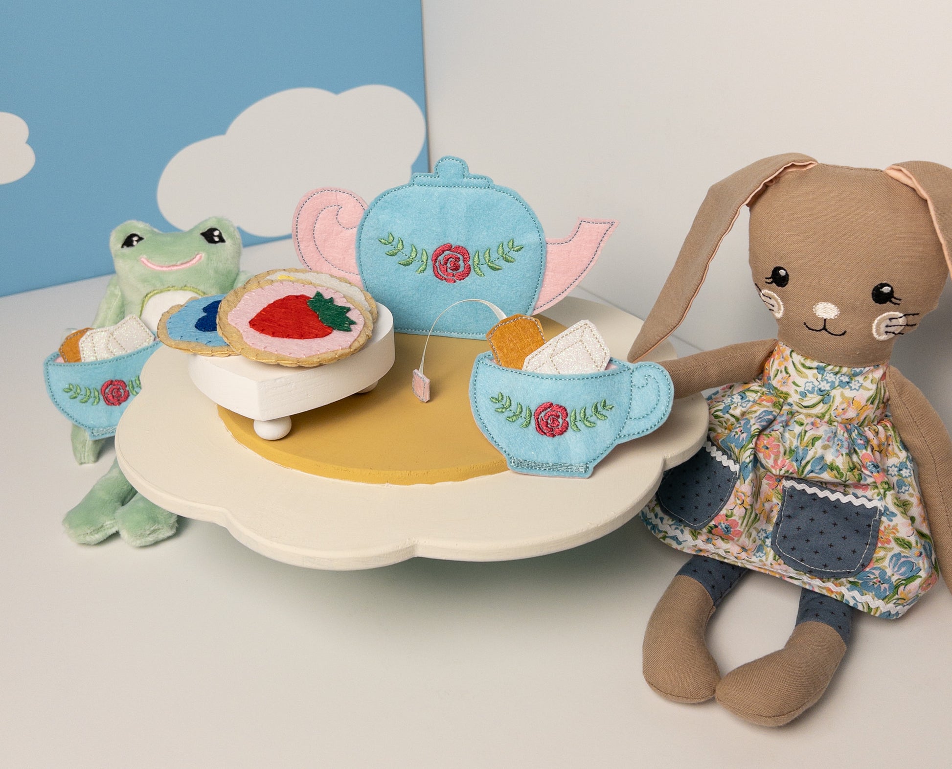 Handmade felt tea set playset on a cloud-shaped table, featuring a blue teapot and cup with rose embroidery, felt cookies, and tea accessories. Accompanied by a stuffed frog, a felt cookie matching game, and a stuffed bunny in a floral dress. Scene set against a background with white clouds on blue sky.