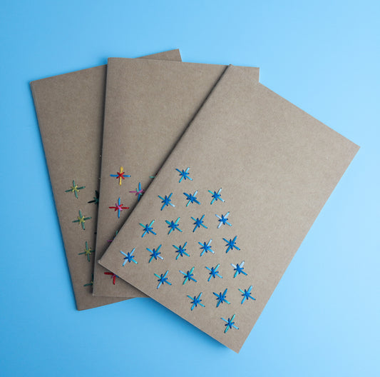 Shows three A5-sized kraft paper notebook journals with colorful hand cross-stitched geometric designs on covers, displayed on blue background