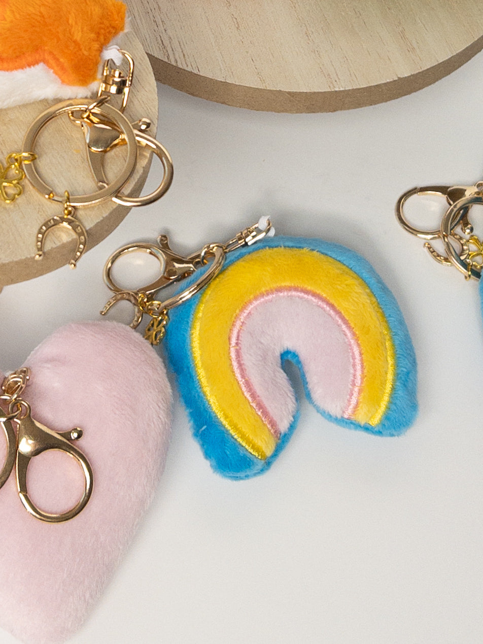 Closeup of plush rainbow bag charm in sky blue, soft pink, and yellow with gold hardware and mini clover accent