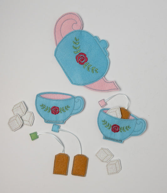 Handcrafted felt tea set on a light background. Set includes a sky-blue teapot with pink spout and lid, two matching teacups, all adorned with red rose and green leaf embroidery. Accessories include tan felt tea bags, white glittery sugar cubes, and pink decorative elements.