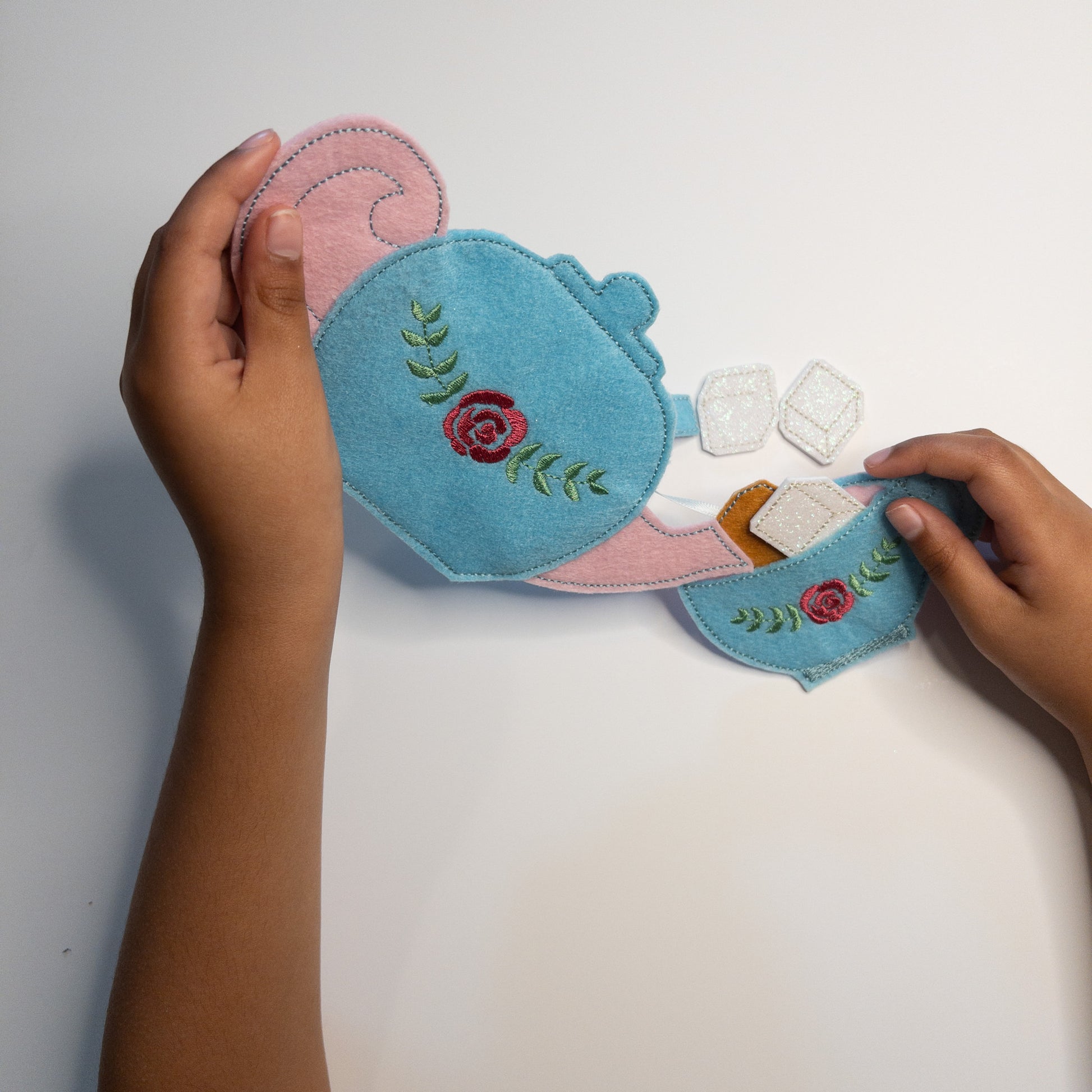 Handmade felt tea set playset featuring a light blue teapot and cups with pink accents and rose embroidery, alongside felt tea bags, sugar cubes, and a child's hands demonstrating play.