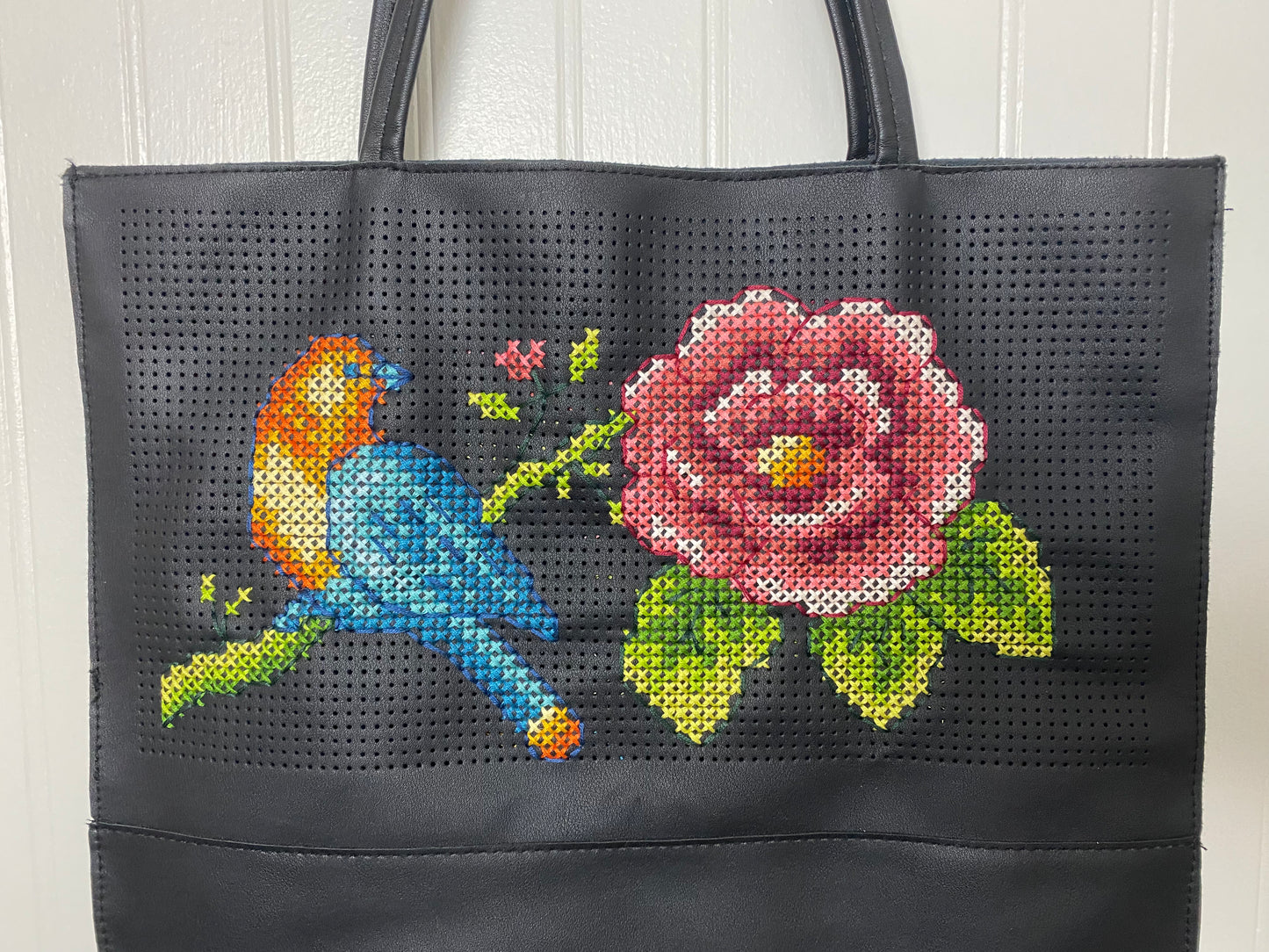 Close-up of intricate hand-stitched cross stitch design featuring vibrant blue bird and red rose on black tote bag fabricand rose.