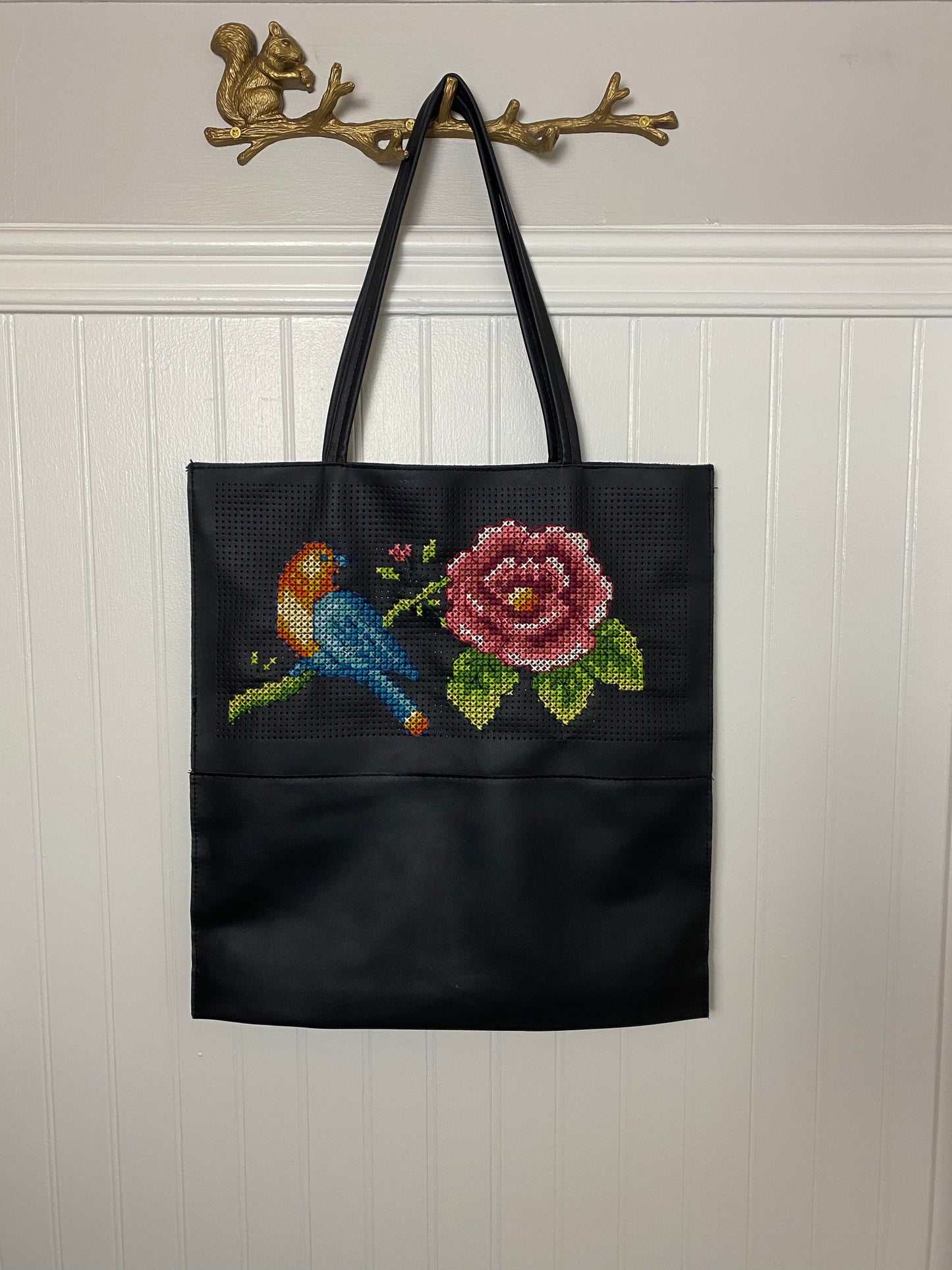 Unique handmade black tote bag with colorful cross-stitched blue bird and pink rose design, hanging on decorative gold squirrel hook and rose.